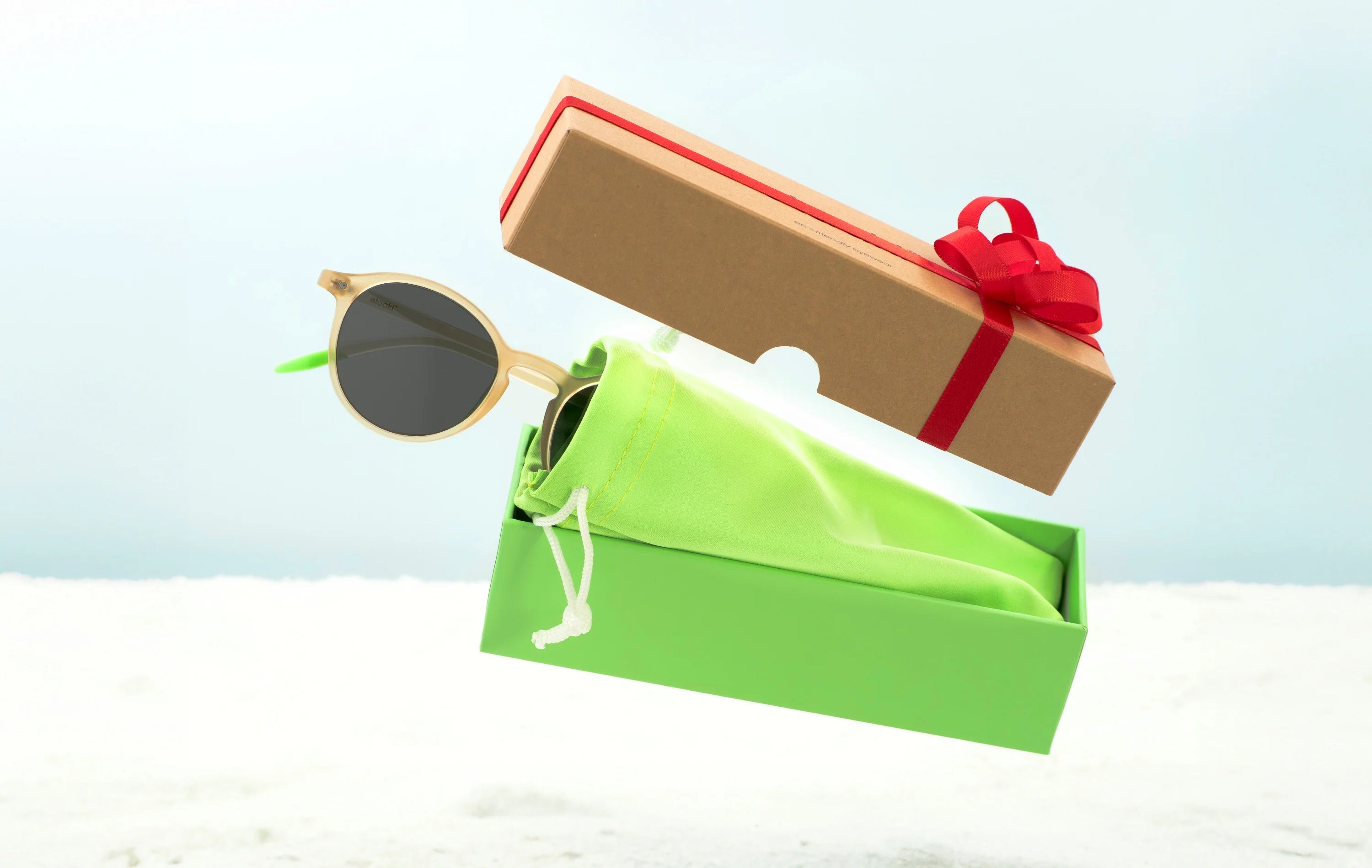 A pair of sunglasses, its cardboard packaging, and its microfiber case, all levitating. The cardboard packaging is adorned with a red Christmas ribbon.