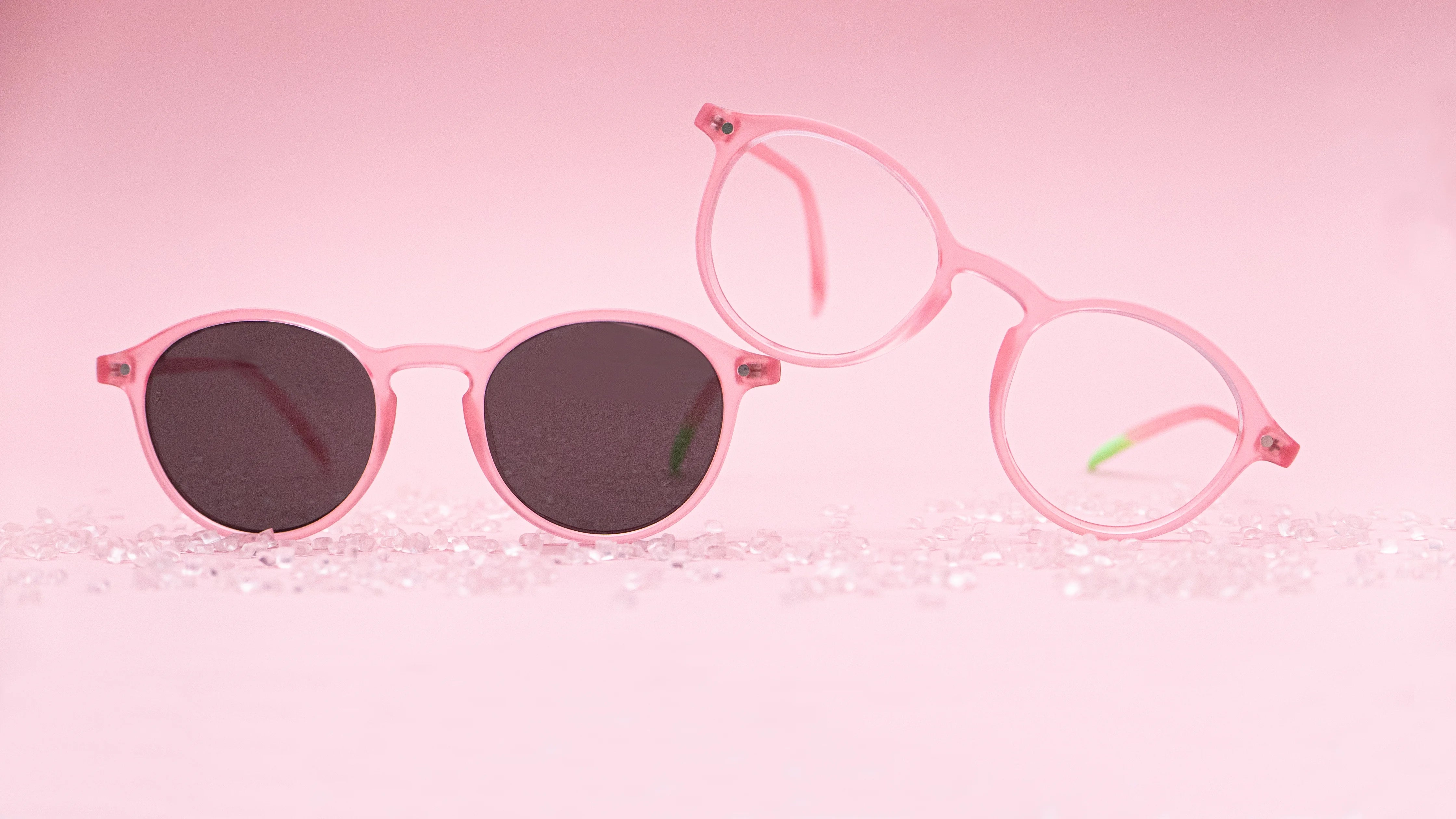 Photograph of 'La Vue En Rose' sunglasses and optical frames. The optical frame overlaps the sunglasses frame. Both frames are surrounded by plastic granules.